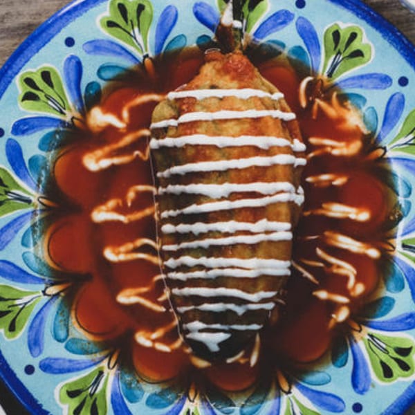 A chile relleno, drizzled with crema and resting in red sauce, on a colorful Mexican plate.