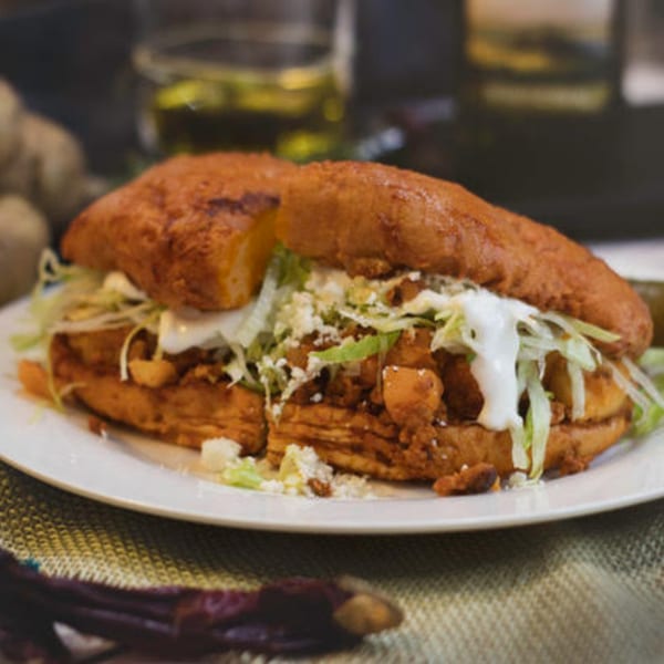 A Mexican pambazo sandwich, filled with meat, lettuce, cheese, and cream, on a white plate.