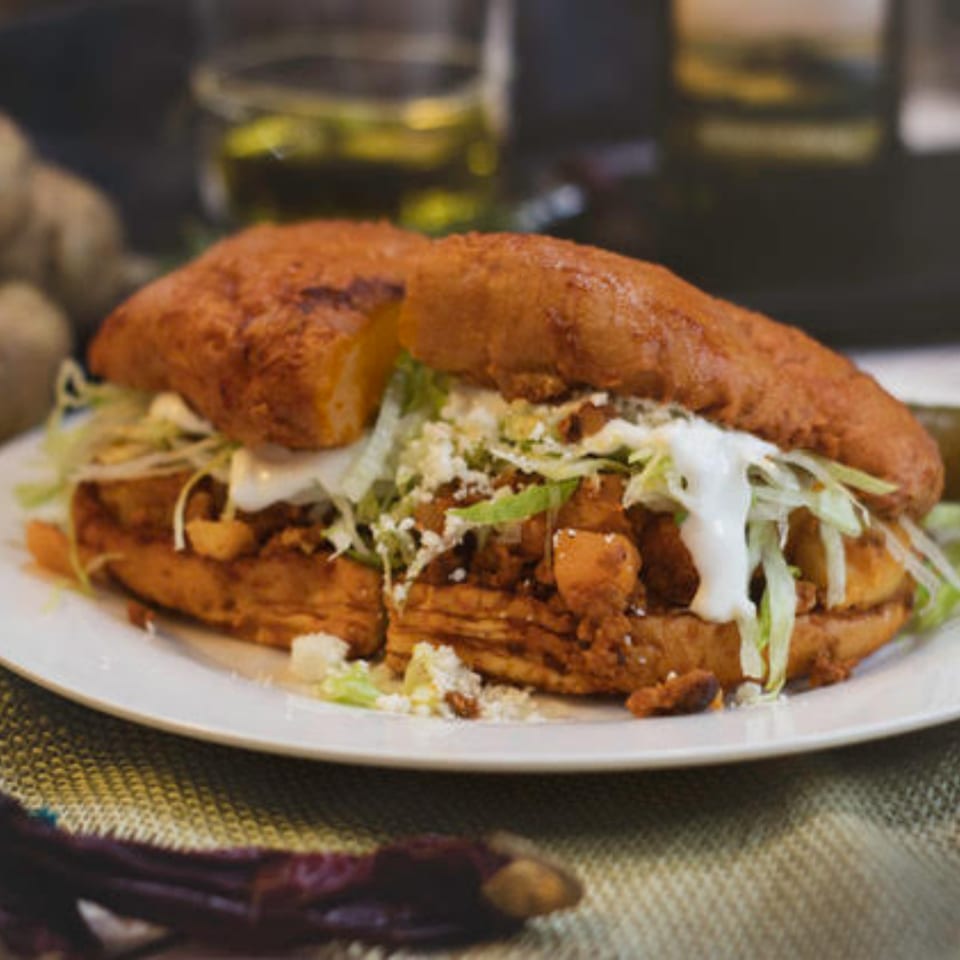 A Mexican pambazo sandwich, filled with meat, lettuce, cheese, and cream, on a white plate.
