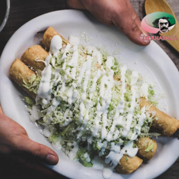 Hands presenting a plate of Taqueria Cuernavaca's golden taquitos topped with shredded lettuce, cheese, and crema.