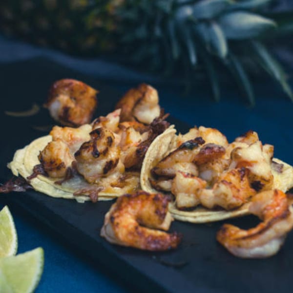 Grilled shrimp tacos with lime wedges on a dark serving board, with a pineapple in the background.
