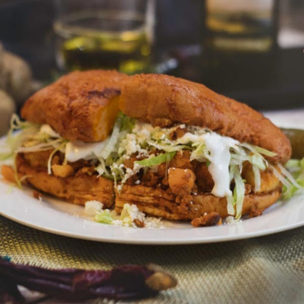 A Mexican pambazo sandwich, filled with meat, lettuce, cheese, and crema, on a white plate.