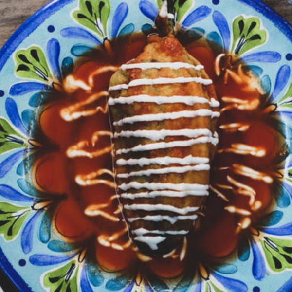 A chile relleno, drizzled with crema and resting in red sauce on a colorful Mexican plate.