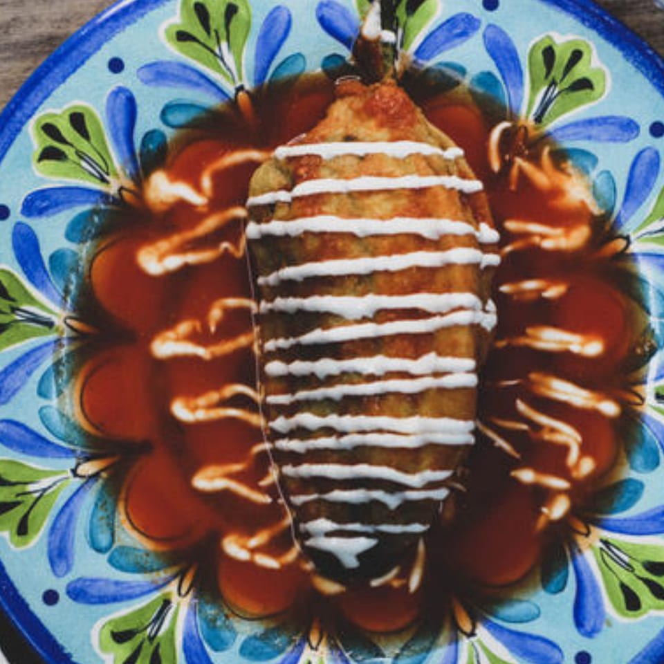 A chile relleno, drizzled with crema and resting in red sauce on a colorful Mexican plate.