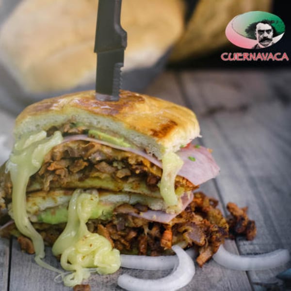 A large Mexican torta sandwich with meat, ham, avocado, and cheese sauce, with a knife stuck in it.