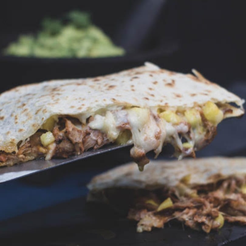 A cheesy quesadilla filled with meat and pineapple, lifted by a spatula, with guacamole in the background.