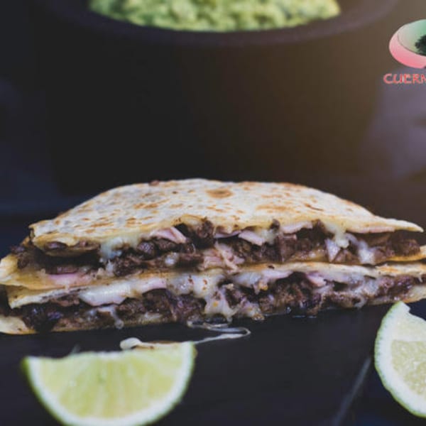 A large, cheesy quesadilla filled with meat and ham, served with lime wedges.