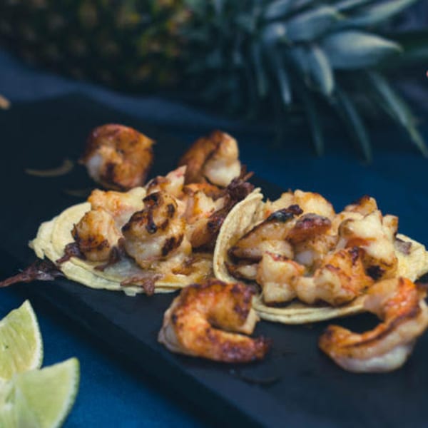Grilled shrimp tacos with lime wedges on a dark platter, with a pineapple in the background.