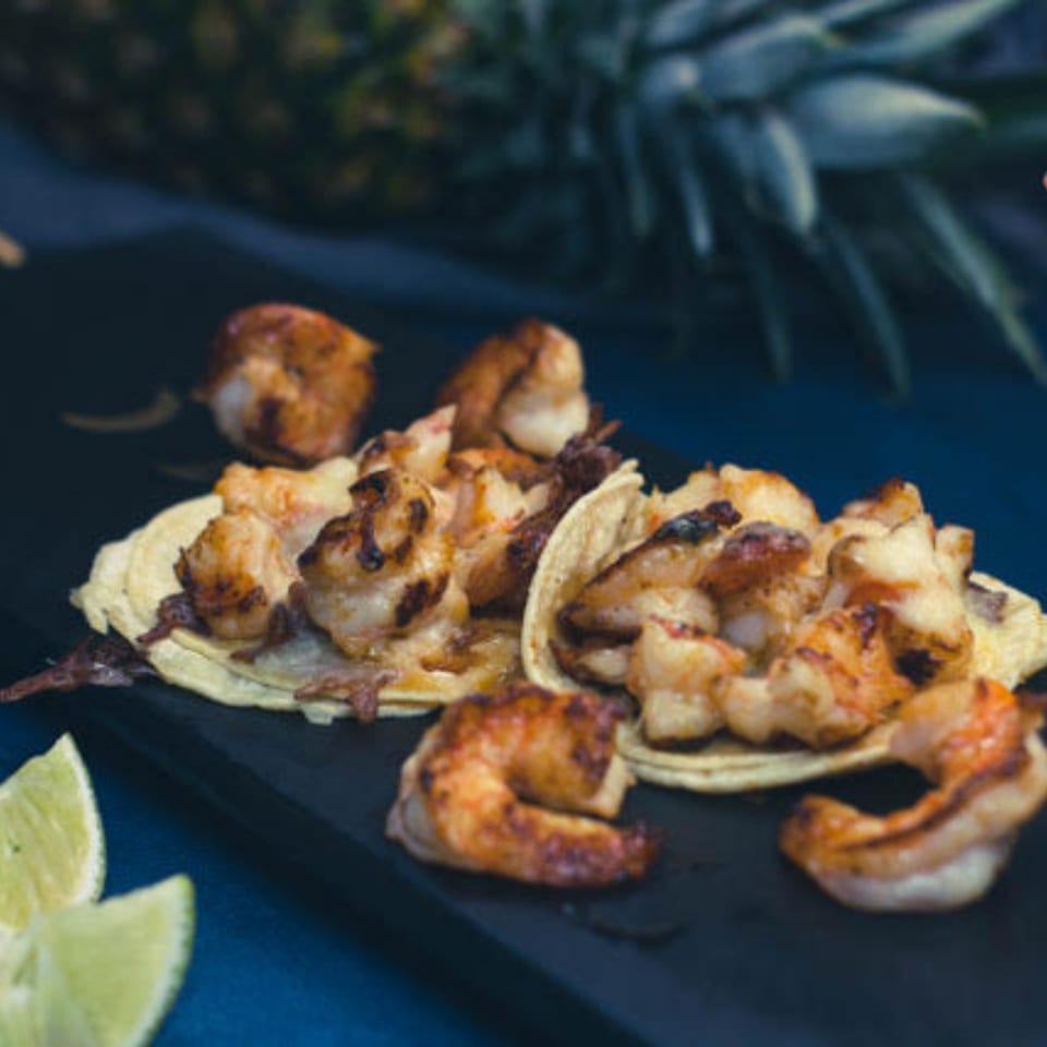 Grilled shrimp tacos with lime wedges on a dark platter, with a pineapple in the background.
