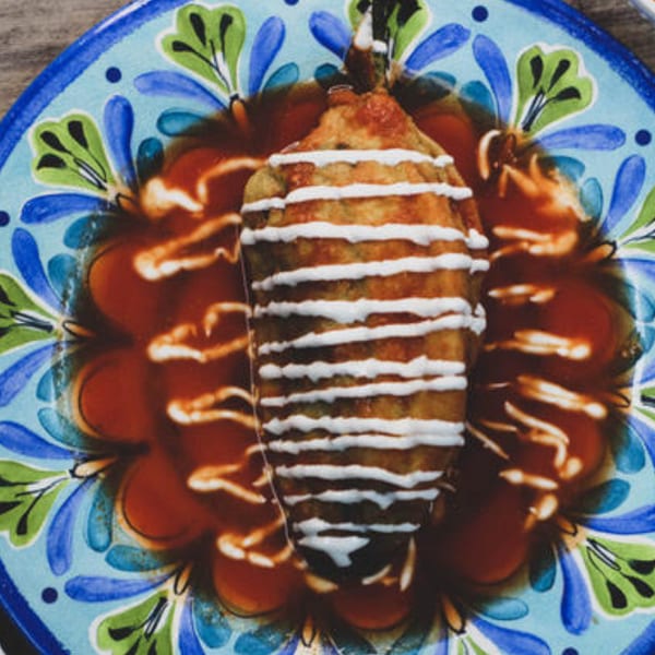 Chili relleno on a colorful plate, drizzled with white cream and surrounded by red sauce.