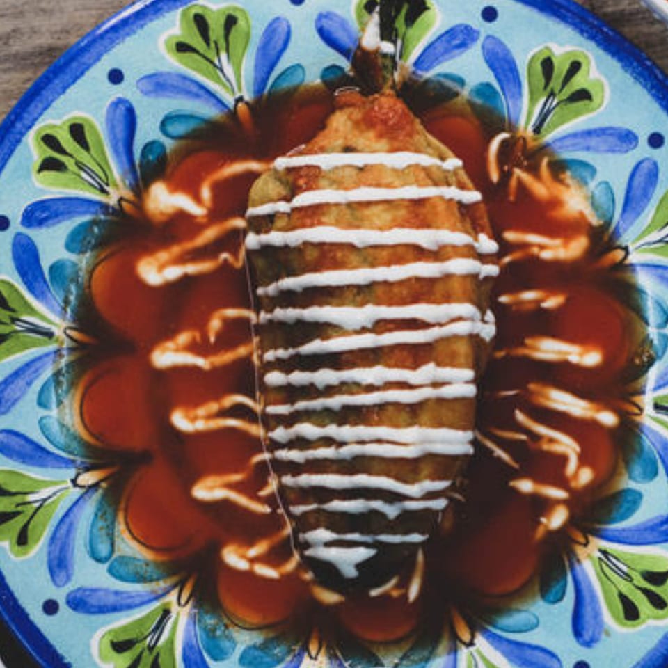 Chili relleno on a colorful plate, drizzled with white cream and surrounded by red sauce.