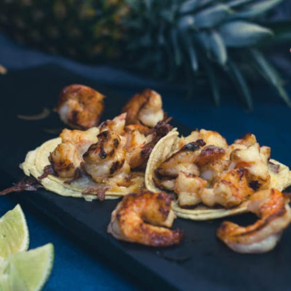 Shrimp tacos with lime wedges and a pineapple in the background on a dark surface.