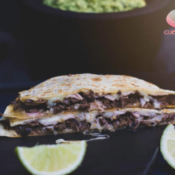 A large, cheesy quesadilla filled with meat, served with lime wedges and guacamole.