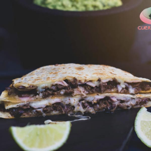 A large, cheesy quesadilla filled with meat, served with lime wedges and guacamole in the background.