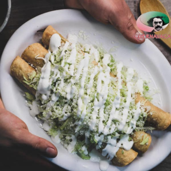 Hands presenting a plate of golden-brown taquitos topped with shredded lettuce, sour cream, and cheese.