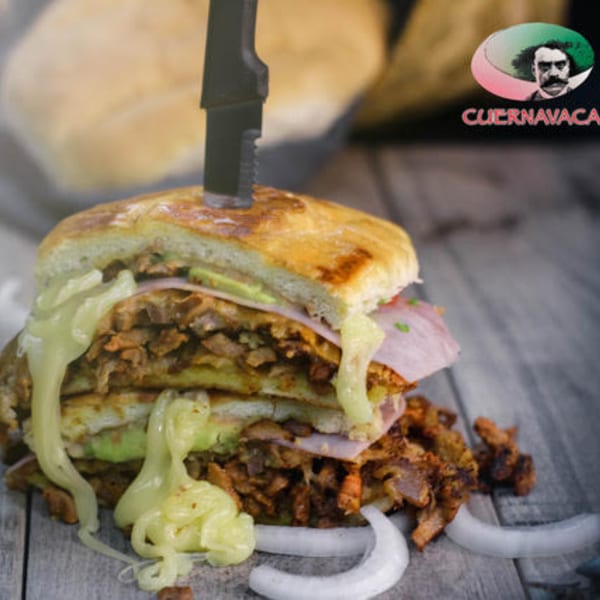 A large Mexican torta sandwich with meat, cheese, avocado, and ham, with a knife stuck in it.