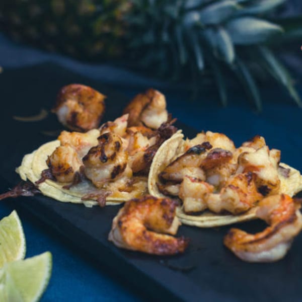 Shrimp tacos with lime wedges on a dark platter, with a pineapple in the background.