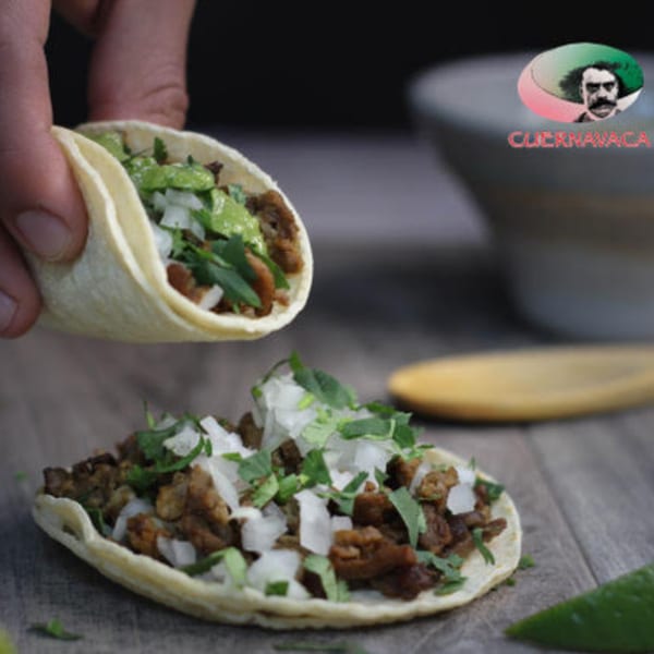Hand holding a taco with meat, guacamole, and cilantro, above another taco on a wooden surface.