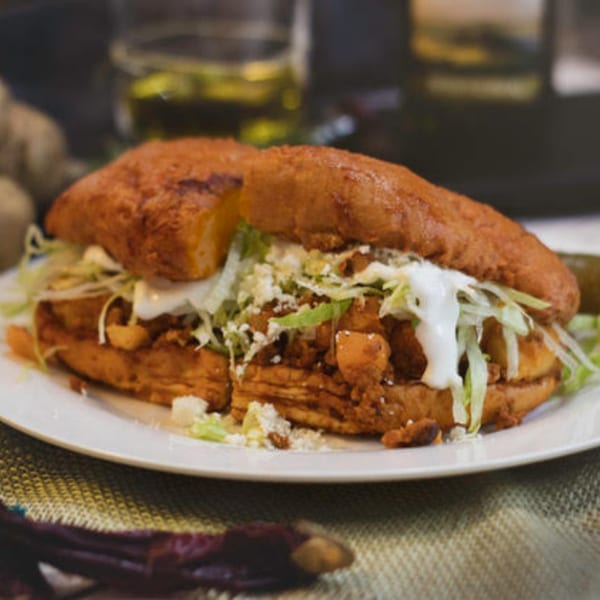 A Mexican pambazo sandwich, filled with meat, lettuce, cheese, and crema, on a white plate.