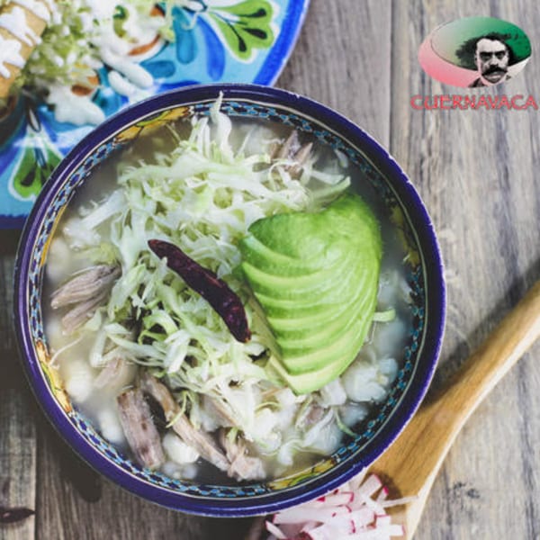A bowl of pozole with shredded cabbage, avocado, and a dried chili on a wooden table.