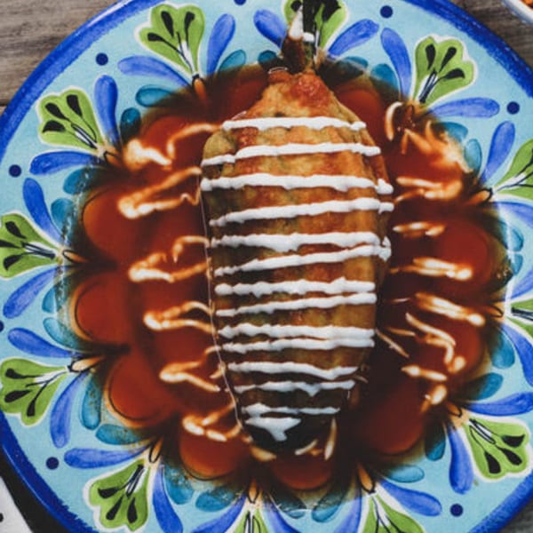 A chile relleno, drizzled with crema, rests in a pool of red sauce on a colorful plate.