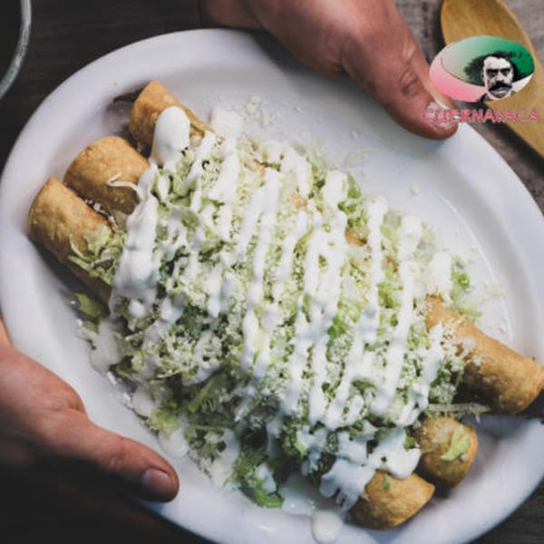 Hands presenting a plate of Taqueria Cuernavaca's golden taquitos topped with lettuce, cheese, and crema.