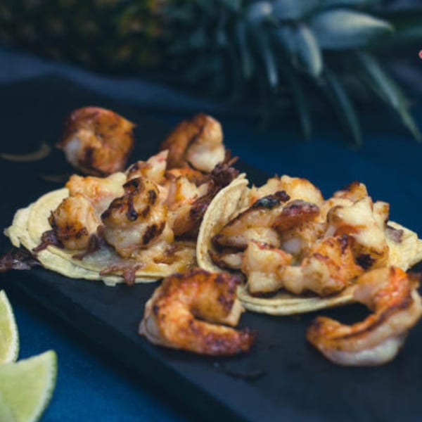Grilled shrimp tacos on corn tortillas with lime wedges and a pineapple in the background.