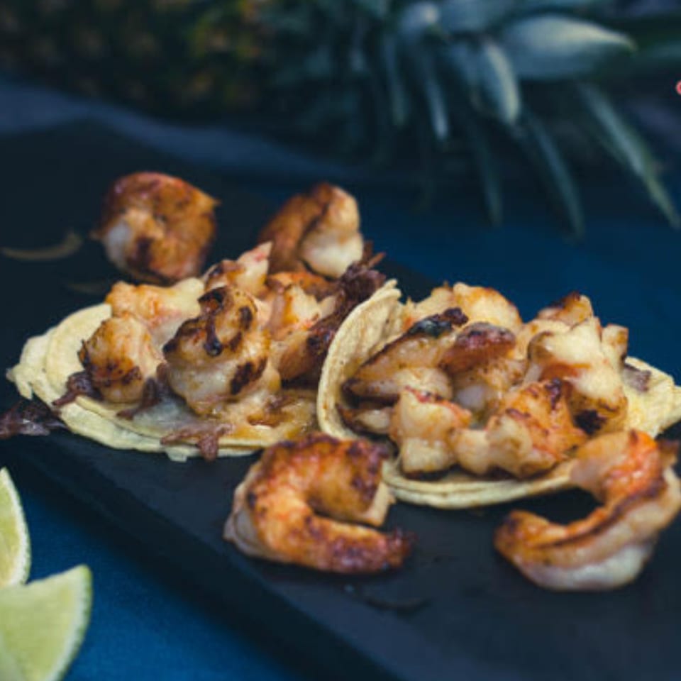 Grilled shrimp tacos on corn tortillas with lime wedges and a pineapple in the background.
