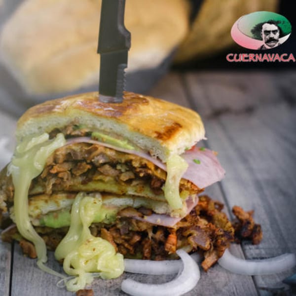 A large Mexican torta sandwich with meat, cheese, and avocado, pierced by a knife.