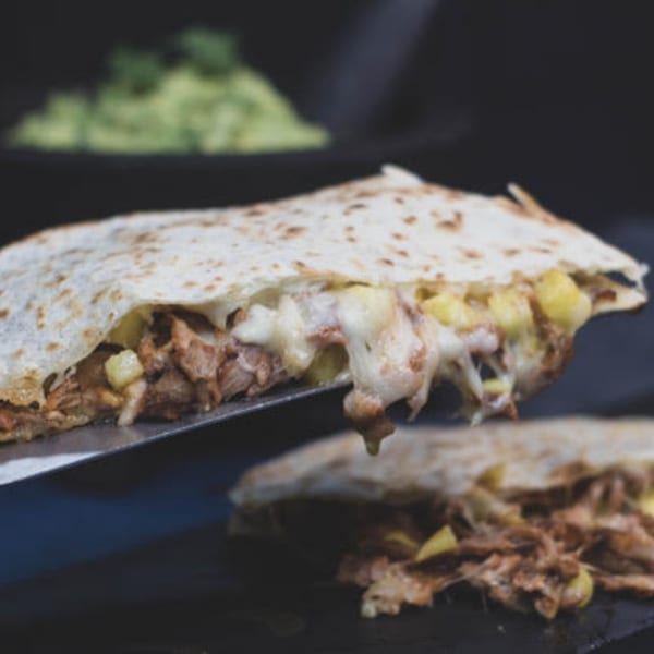 A quesadilla filled with melted cheese, shredded meat, and pineapple, with guacamole in the background.