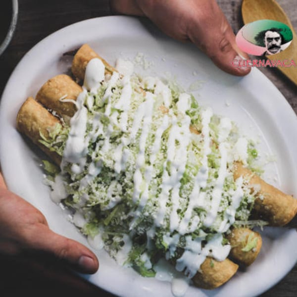 Hands presenting a plate of Taqueria Cuernavaca's golden flautas topped with lettuce, cheese, and crema.