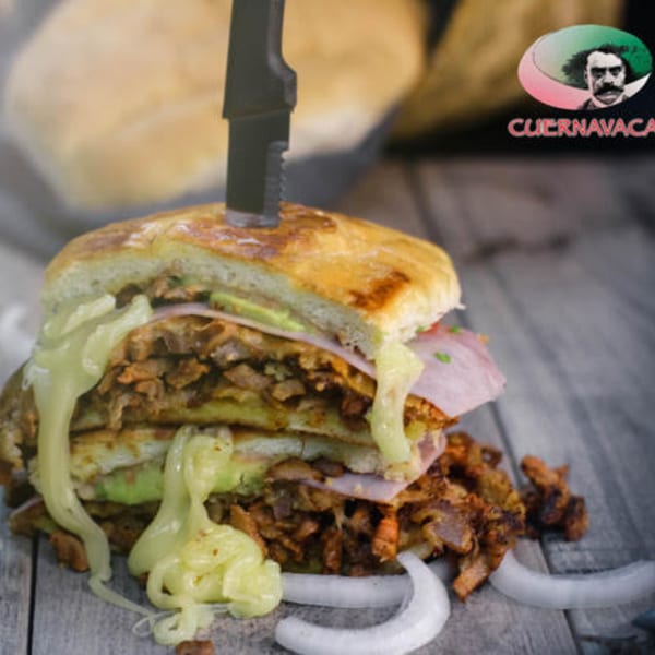A large Mexican torta sandwich with meat, cheese, avocado, and ham, pierced by a knife.